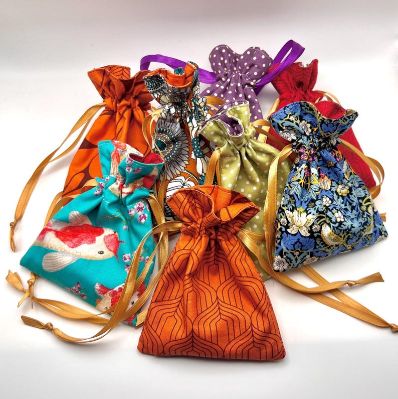 May include: A collection of colorful drawstring pouches in various patterns. The pouches feature designs such as koi fish, floral prints, polka dots, and geometric shapes. Each pouch is tied with a contrasting ribbon or gold cord.