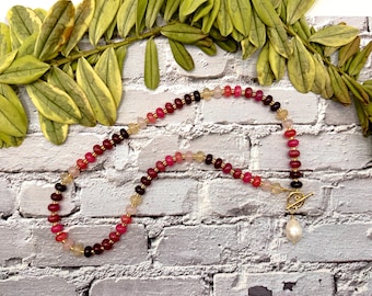 May include: A beaded necklace with a variety of colorful gemstones, including red, pink, and clear beads. The necklace has a gold clasp and a pear-shaped pearl pendant. The necklace is displayed on a white brick background with green foliage.