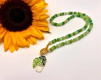 May include: A green beaded necklace with a gold clasp and a pendant featuring a plant design. The necklace is displayed next to a bright yellow sunflower with a dark brown center. The necklace is a unique piece of jewelry.