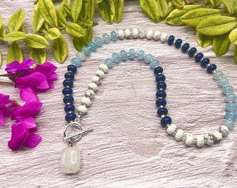 May include: A beaded necklace featuring a mix of blue, white, and navy beads. The necklace has a silver toggle clasp and a light-colored, rectangular pendant. The beads are arranged in a gradient pattern, with a mix of round and irregular shapes.