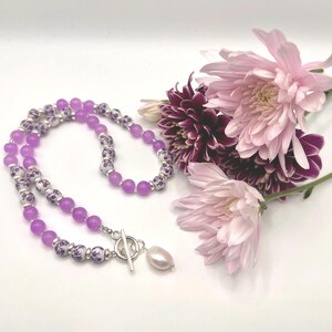 May include: A necklace with lavender beads and decorative white and purple beads. The necklace has a silver clasp and a single pearl pendant. Pink and purple flowers are arranged near the necklace on a white surface.