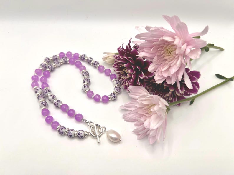 May include: A necklace with lavender beads and decorative white and purple beads. The necklace has a silver clasp and a single pearl pendant. Pink and purple flowers are arranged near the necklace on a white surface.