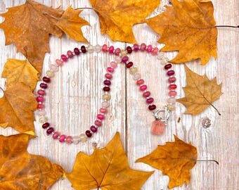 May include: A beaded necklace with a variety of pink and red stones, including a square-shaped pendant. The necklace is fastened with a silver clasp. The background features autumn leaves on a wooden surface.