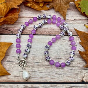 May include: A necklace featuring alternating purple and white floral beads, with a large white pearl pendant. The necklace is displayed on a wooden surface with autumn leaves.