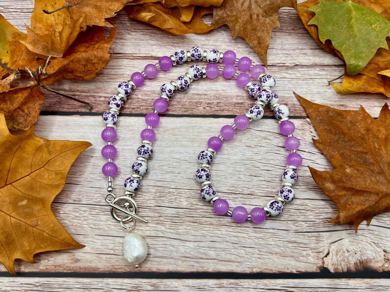 May include: A necklace featuring alternating purple and white floral beads, with a large white pearl pendant. The necklace is displayed on a wooden surface with autumn leaves.