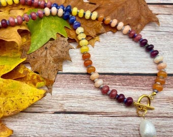 May include: A colorful beaded necklace with a gold clasp and a white teardrop-shaped pendant. The necklace features a gradient of colors, including yellow, orange, pink, and purple beads. The necklace is displayed on a wooden surface with autumn leaves.