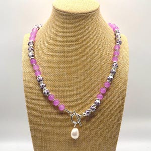 May include: A necklace with a toggle clasp and a dangling white pearl pendant. The necklace features alternating light purple and patterned beads. The patterned beads are white with a dark purple floral design. The necklace is displayed on a tan jewelry bust.