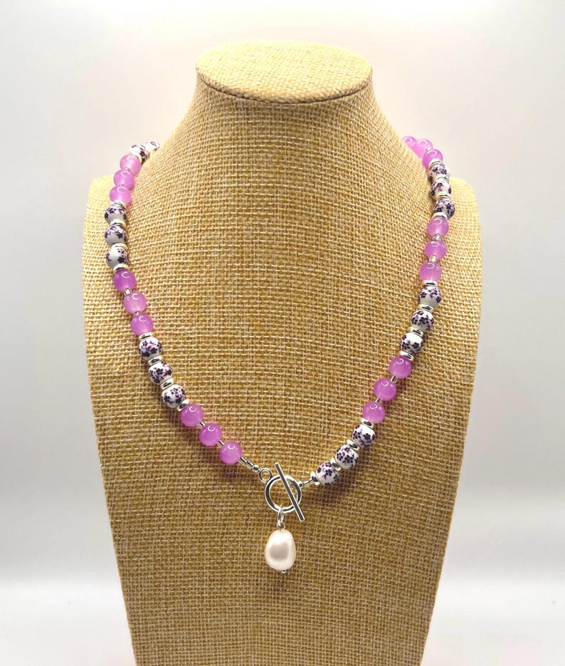 May include: A necklace with a toggle clasp and a dangling white pearl pendant. The necklace features alternating light purple and patterned beads. The patterned beads are white with a dark purple floral design. The necklace is displayed on a tan jewelry bust.