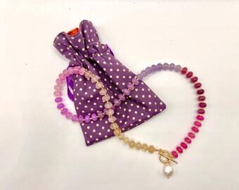 May include: A multi-colored beaded necklace with a gold clasp and a pearl pendant. The necklace is displayed next to a purple polka-dot drawstring bag. The beads transition from pink to purple to yellow and red.
