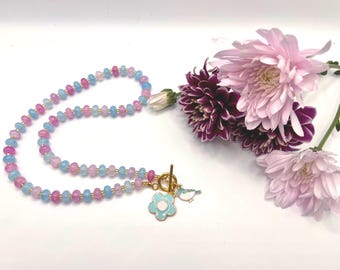 May include: A delicate necklace featuring pink and blue gemstone beads strung together with gold-colored accents. The necklace has a gold clasp and a light blue flower charm with a small bird pendant. Pink and purple flowers are in the background.