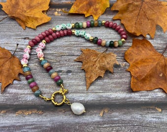 May include: A colorful beaded necklace with a gold clasp and a white pearl pendant. The necklace features a variety of beads in shades of pink, green, blue, and gold. The necklace is displayed on a wooden surface with autumn leaves.
