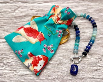 May include: A turquoise drawstring pouch with a koi fish print and a beaded necklace. The necklace features blue and clear beads with a silver clasp and a dark blue pendant.