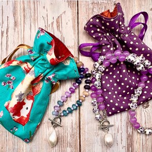 May include: Two beaded necklaces with pearl pendants and toggle clasps rest beside drawstring pouches. One pouch is teal with a koi fish pattern, the other is purple with white polka dots. The necklaces feature purple, white, and blue beads.