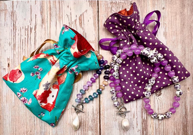 May include: Two beaded necklaces with pearl pendants and toggle clasps rest beside drawstring pouches. One pouch is teal with a koi fish pattern, the other is purple with white polka dots. The necklaces feature purple, white, and blue beads.