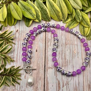 May include: A necklace featuring alternating purple and white floral patterned beads. The necklace has a silver clasp and a white pearl pendant. The necklace is displayed on a wooden surface with green foliage in the background.