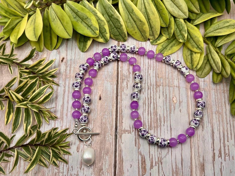 May include: A necklace featuring alternating purple and white floral patterned beads. The necklace has a silver clasp and a white pearl pendant. The necklace is displayed on a wooden surface with green foliage in the background.
