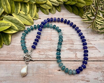 May include: A beaded necklace with a toggle clasp and a teardrop-shaped pearl pendant. The necklace features a gradient of blue and teal beads. The necklace is displayed on a light wood surface with green foliage in the background.