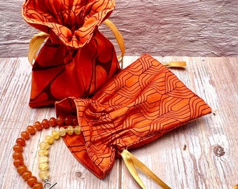May include: Two orange fabric drawstring bags with a geometric pattern and gold ribbon ties. A beaded necklace with a pearl pendant is partially visible, emerging from one bag. The bags are on a wooden surface.
