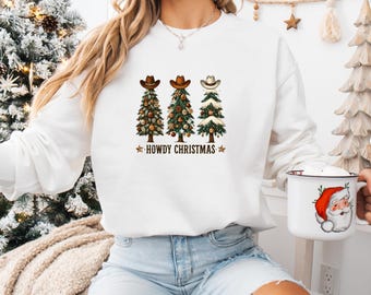May include: White crew neck sweatshirt with a festive design. The design features three Christmas trees topped with cowboy hats, decorated with ornaments, and the text "HOWDY CHRISTMAS". The sweatshirt has long sleeves.