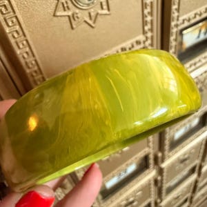 May include: A wide, lime green bangle bracelet with a marbled effect. The chunky, rounded bracelet is held up against a background of gold-colored mailboxes. The bracelet's surface has a glossy finish.