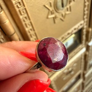 May include: A silver ring featuring a large, oval-cut ruby gemstone. The ruby is a deep red color with visible facets. The ring's band is a simple, polished silver. The background is a gold-colored surface with decorative details.