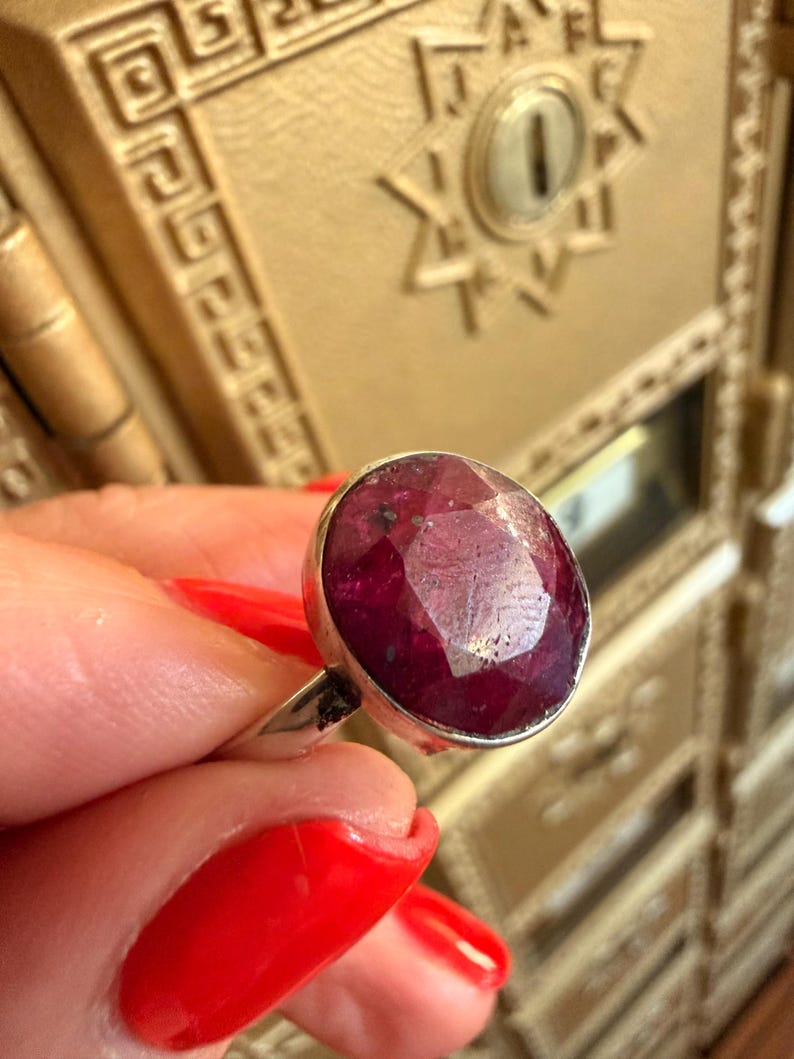 May include: A silver ring featuring a large, oval-cut ruby gemstone. The ruby is a deep red color with visible facets. The ring's band is a simple, polished silver. The background is a gold-colored surface with decorative details.