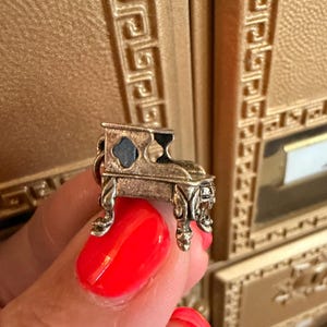May include: A small, metallic charm shaped like a miniature piano with ornate, curved legs and a dark, recessed interior. The charm features a detailed, textured surface and a small loop at the top for attachment to a bracelet or necklace chain.