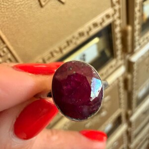 May include: A silver ring featuring a large, oval-cut ruby gemstone. The ruby is a deep, rich red color with facets that catch the light. The ring has a simple, elegant design, showcasing the beauty of the gemstone.