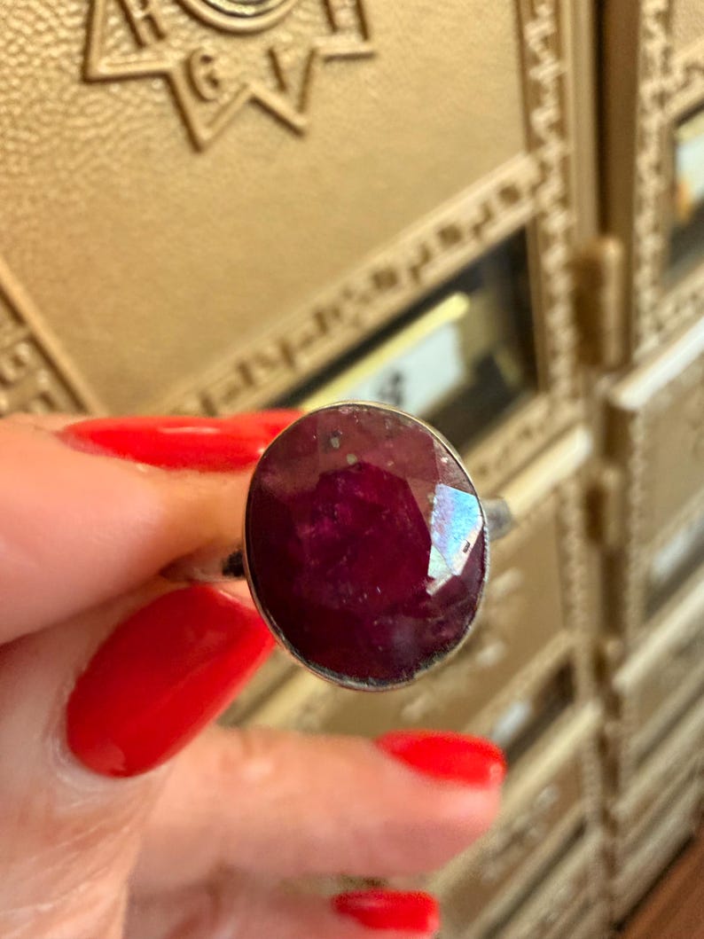 May include: A silver ring featuring a large, oval-cut ruby gemstone. The ruby is a deep, rich red color with facets that catch the light. The ring has a simple, elegant design, showcasing the beauty of the gemstone.