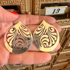May include: A pair of gold-toned metal earrings with a teardrop shape and an intricate cut-out filigree pattern featuring swirling, organic lines. The earrings have a smooth, reflective surface and a pointed top section with a rounded base.