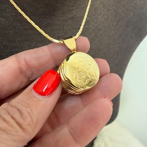 May include: A gold-colored locket necklace with a floral design on the front. The locket is round and hangs from a delicate gold chain. The necklace is held by a hand with red nail polish.