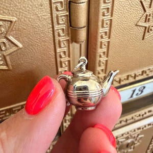 May include: A small, silver-colored teapot charm with a handle, spout, and lid. The charm is held in front of a gold-colored background. The number "13" is visible in the background.