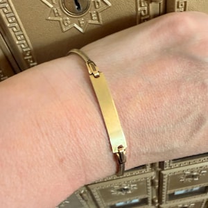May include: A gold-toned identification bracelet featuring a smooth, rectangular metal plate centered on a flexible, textured chain link band. The bracelet has a polished finish and a secure clasp mechanism, designed for a comfortable fit around the wrist.