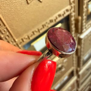 May include: A silver ring featuring a large, oval-cut ruby gemstone. The ruby is a deep red color and is set in a simple silver band. The ring is held up against a gold-colored background.