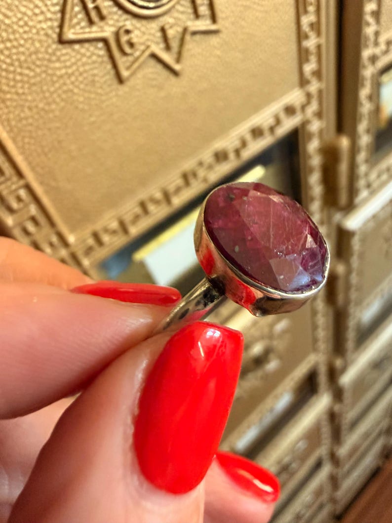 May include: A silver ring featuring a large, oval-cut ruby gemstone. The ruby is a deep red color and is set in a simple silver band. The ring is held up against a gold-colored background.
