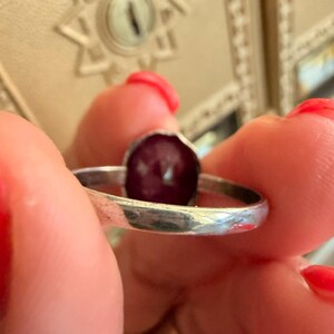 May include: A silver ring with a dark red, oval-shaped stone. The ring is held between fingers with red nail polish. The ring's band is thin and has a slightly hammered texture. The stone is set in a simple bezel.