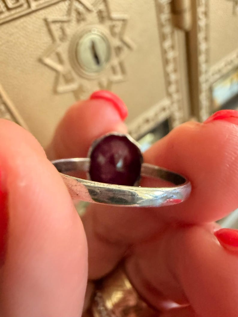 May include: A silver ring with a dark red, oval-shaped stone. The ring is held between fingers with red nail polish. The ring's band is thin and has a slightly hammered texture. The stone is set in a simple bezel.