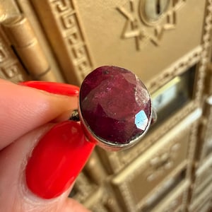 May include: A silver ring featuring a large, oval-cut ruby gemstone. The ruby is a deep, rich red color with facets that catch the light. The ring is held up against a gold-colored background.
