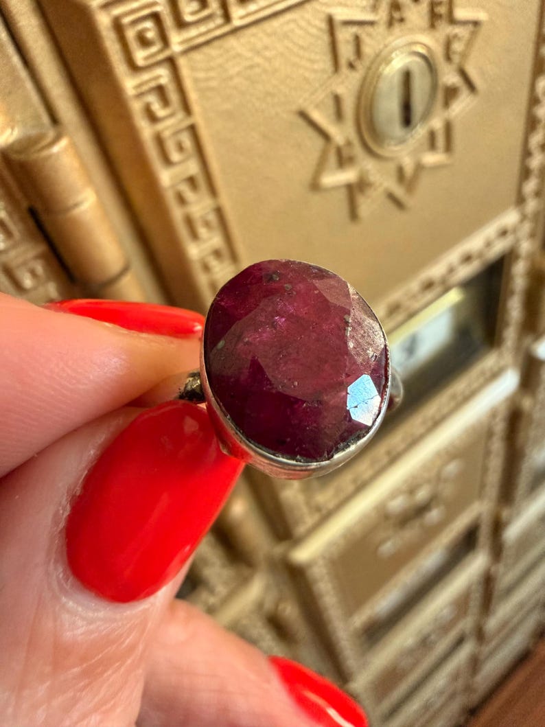 May include: A silver ring featuring a large, oval-cut ruby gemstone. The ruby is a deep, rich red color with facets that catch the light. The ring is held up against a gold-colored background.