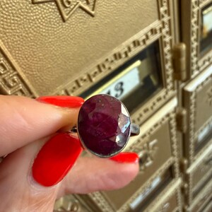 May include: A silver ring featuring a large, faceted oval ruby gemstone. The ruby is a deep, rich red color. The ring is held up against a gold-colored background.
