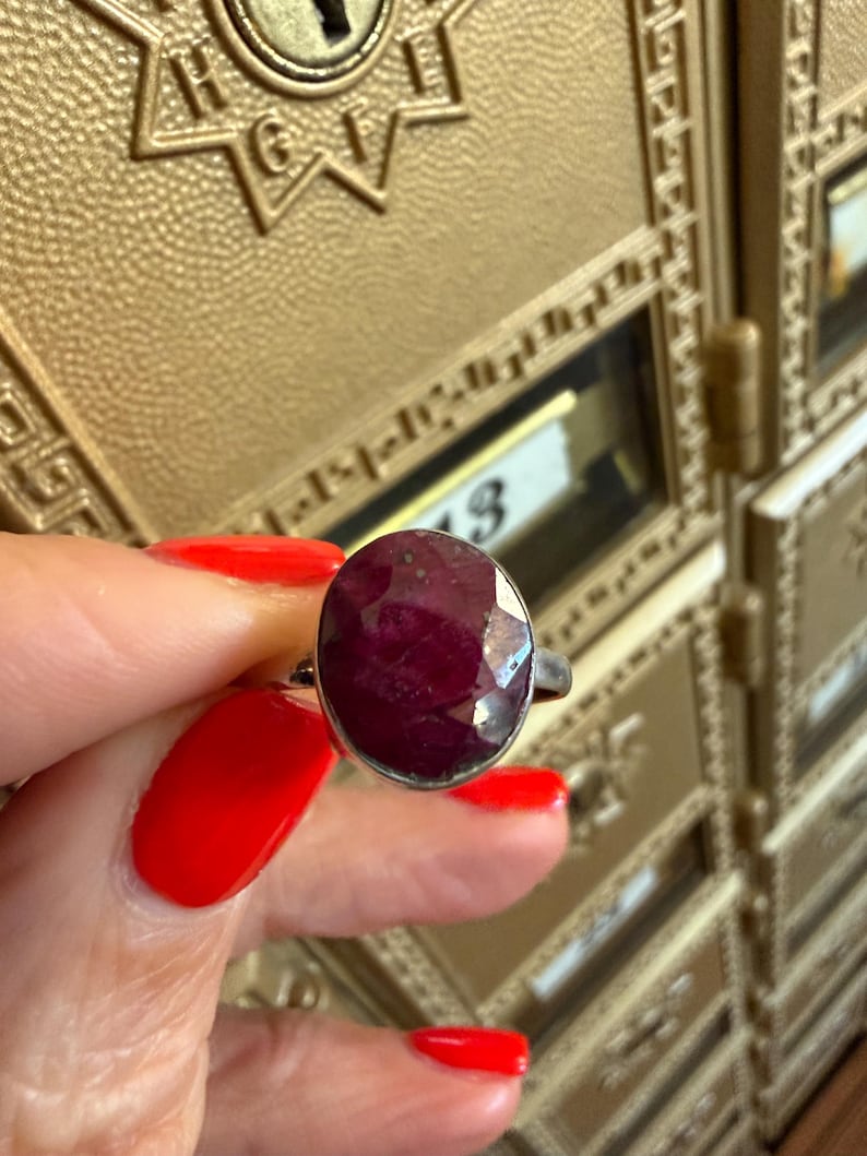 May include: A silver ring featuring a large, faceted oval ruby gemstone. The ruby is a deep, rich red color. The ring is held up against a gold-colored background.