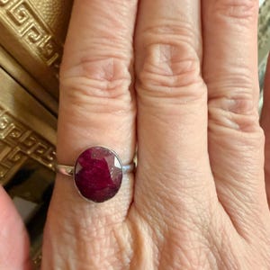 May include: A silver ring featuring a large, oval-cut ruby gemstone. The ruby has a deep red color and is set in a simple, polished silver band. The ring is shown on a person's finger.