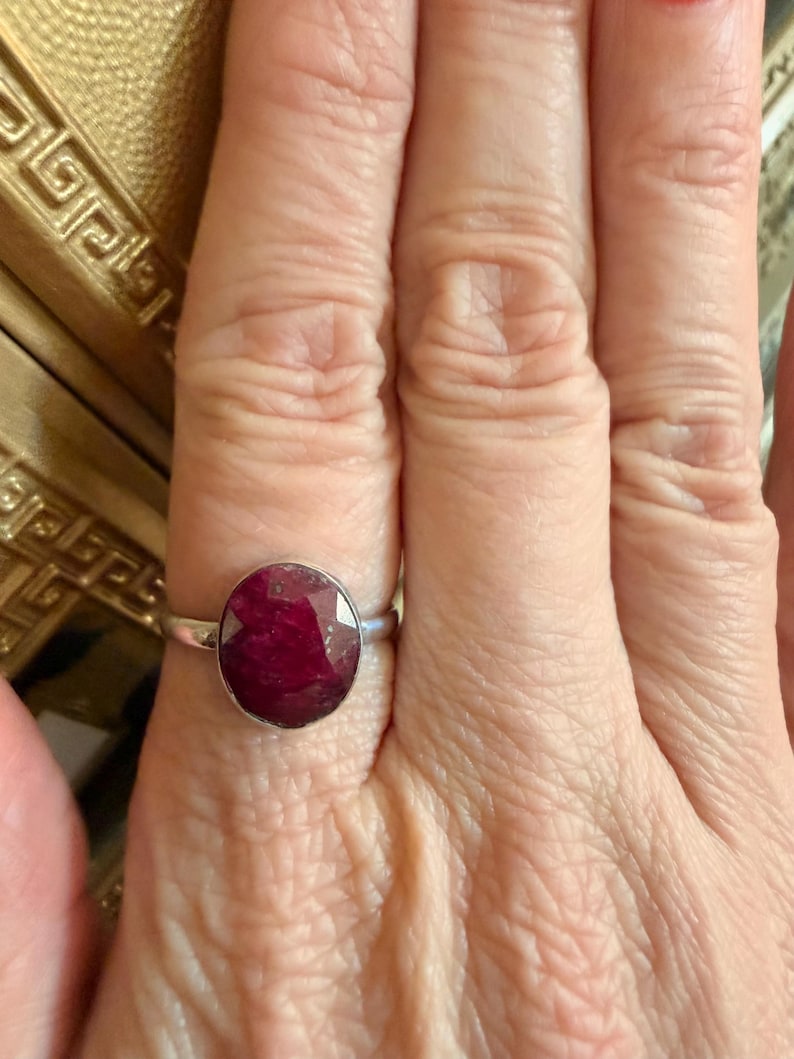 May include: A silver ring featuring a large, oval-cut ruby gemstone. The ruby has a deep red color and is set in a simple, polished silver band. The ring is shown on a person's finger.