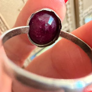 May include: A silver ring featuring a faceted, deep red gemstone. The gemstone is round and set in a bezel setting. The ring band is a simple, polished silver. The ring is held up by a person's fingers.