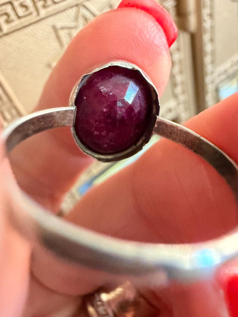 May include: A silver ring featuring a faceted, deep red gemstone. The gemstone is round and set in a bezel setting. The ring band is a simple, polished silver. The ring is held up by a person's fingers.