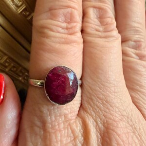 May include: A silver ring featuring a large, oval-cut ruby gemstone. The ruby has a deep red color with visible inclusions. The ring is set on a woman's finger, showcasing the jewelry's design and color.