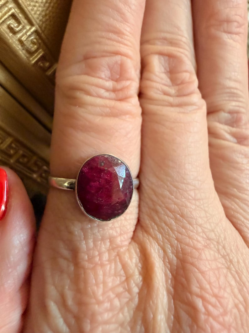 May include: A silver ring featuring a large, oval-cut ruby gemstone. The ruby has a deep red color with visible inclusions. The ring is set on a woman's finger, showcasing the jewelry's design and color.