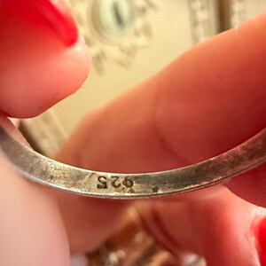 May include: Close-up of a silver ring with the marking "925" visible. The ring is held between fingers with red nail polish. The background is blurred, showing a glimpse of a decorative surface.