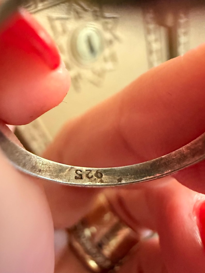 May include: Close-up of a silver ring with the marking "925" visible. The ring is held between fingers with red nail polish. The background is blurred, showing a glimpse of a decorative surface.