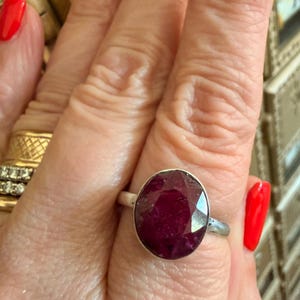May include: A silver ring featuring a large, oval-cut ruby gemstone. The ruby is a deep red color and set in a simple silver band. The ring is shown on a hand with red nail polish.